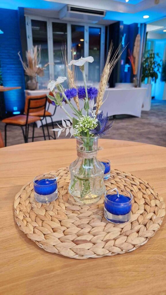 Centre de table, vase en verre, fleurs blanches, fleurs bleues, épi de blé, bougies bleues sur un dessous d'assiette en osier pour un mariage guinguette avec Nature Event